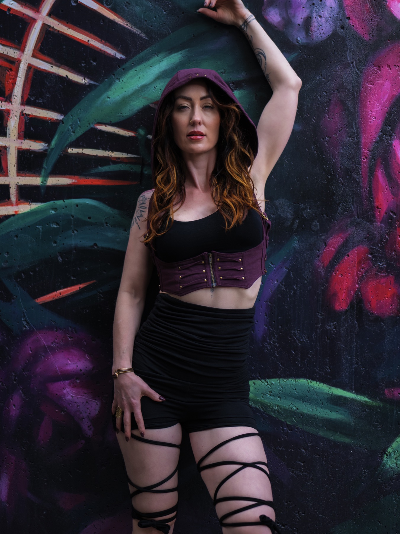 A model faces the camera with one arm raised, wearing a plum hooded crop top, black skirt, and wrapped leg straps against a dark teal and magenta mural.
