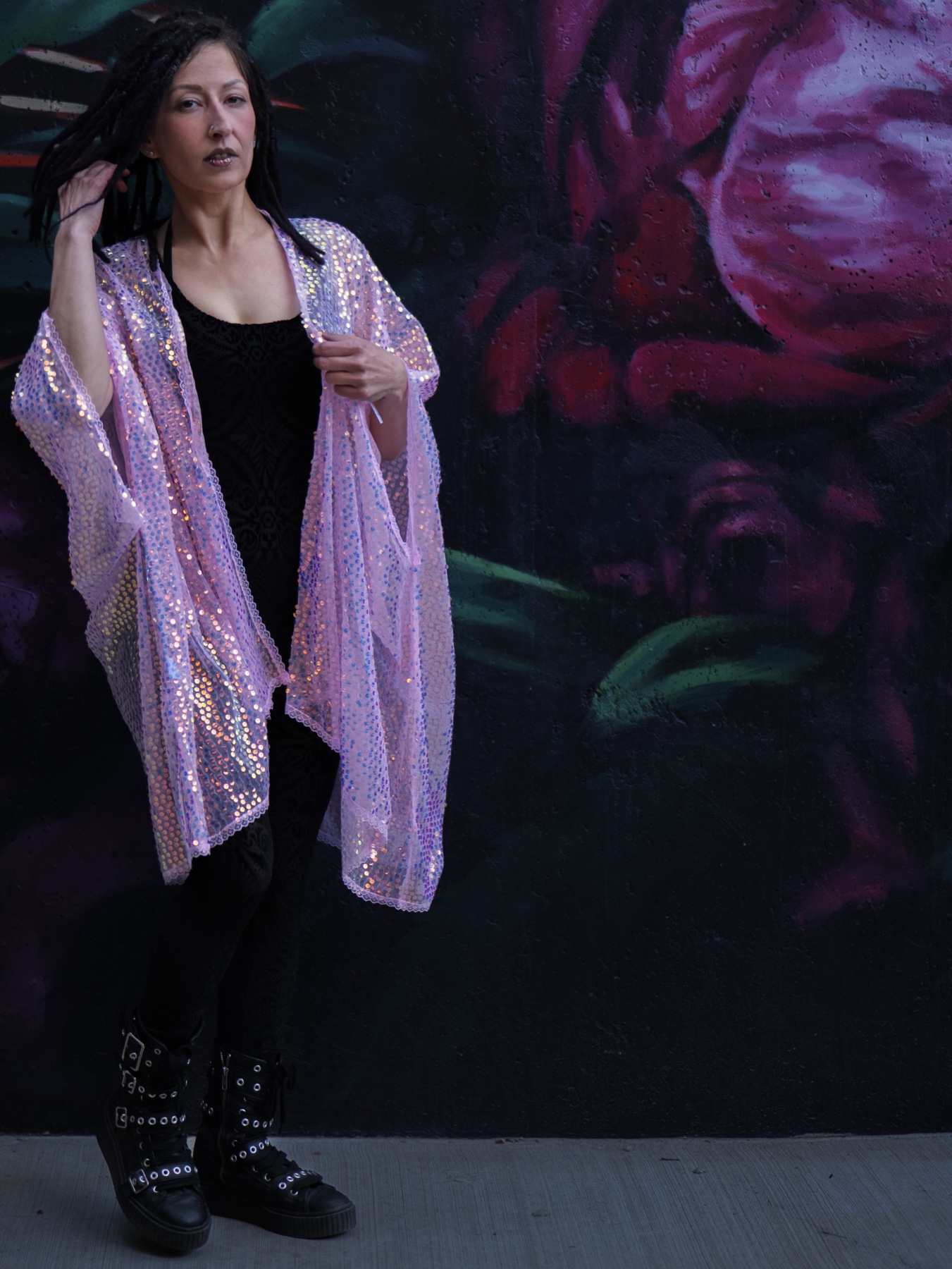 A model stands against a dark floral mural wearing a black outfit with a sheer iridescent sequin layer, showing how texture and reflective fabric can become a strong retail styling image.